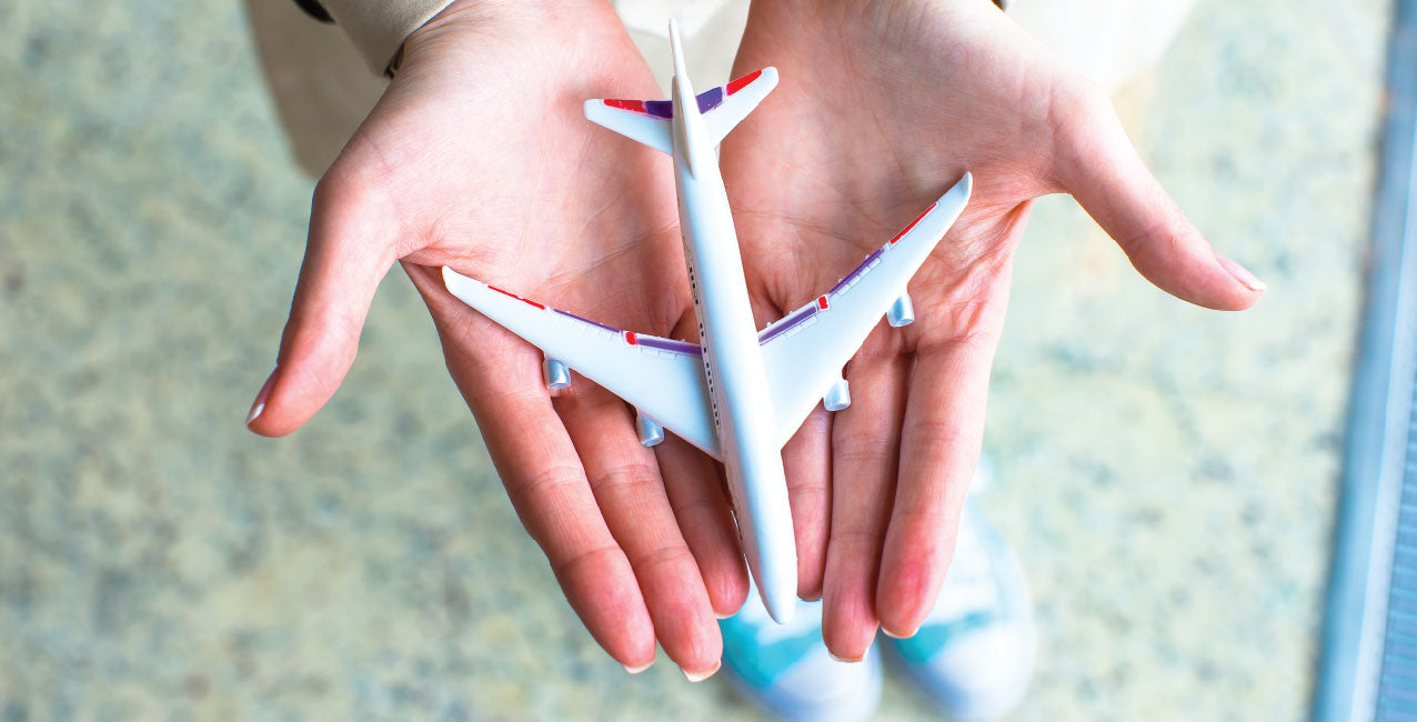 A white woman with French tipped nails holds a small model plane in her hands. You can not see her face and can just see her blue sneakers against the tan floor. 