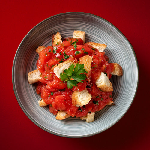 Photo of bruschetta in a bowl over bread. The bowl is grey and placed on a red background.