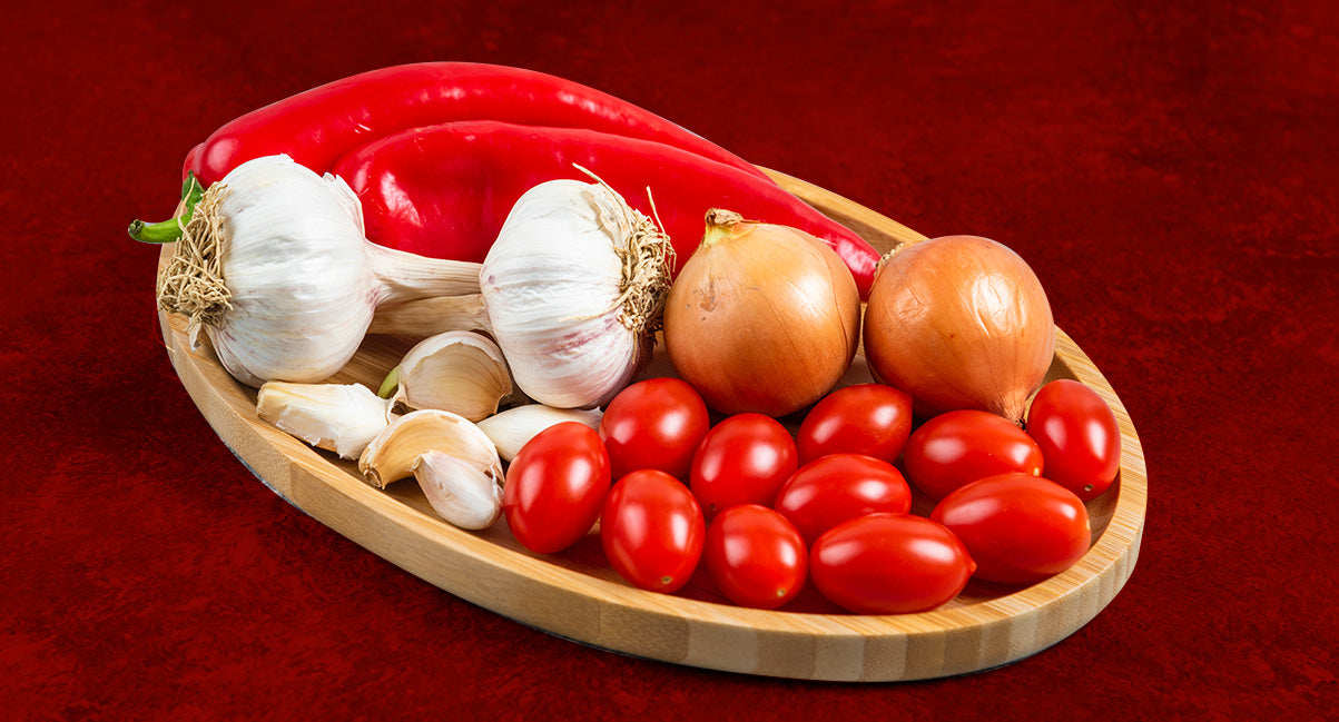 A cutting board with garlic, onions, tomatoes and peppers on a red background.