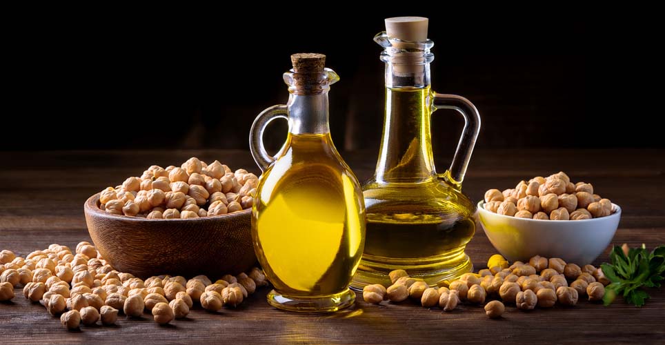 A photo of two decanters of olive oil next to two overflowing bowls of chickpeas. They're on a wooden surface and in front of a black background.