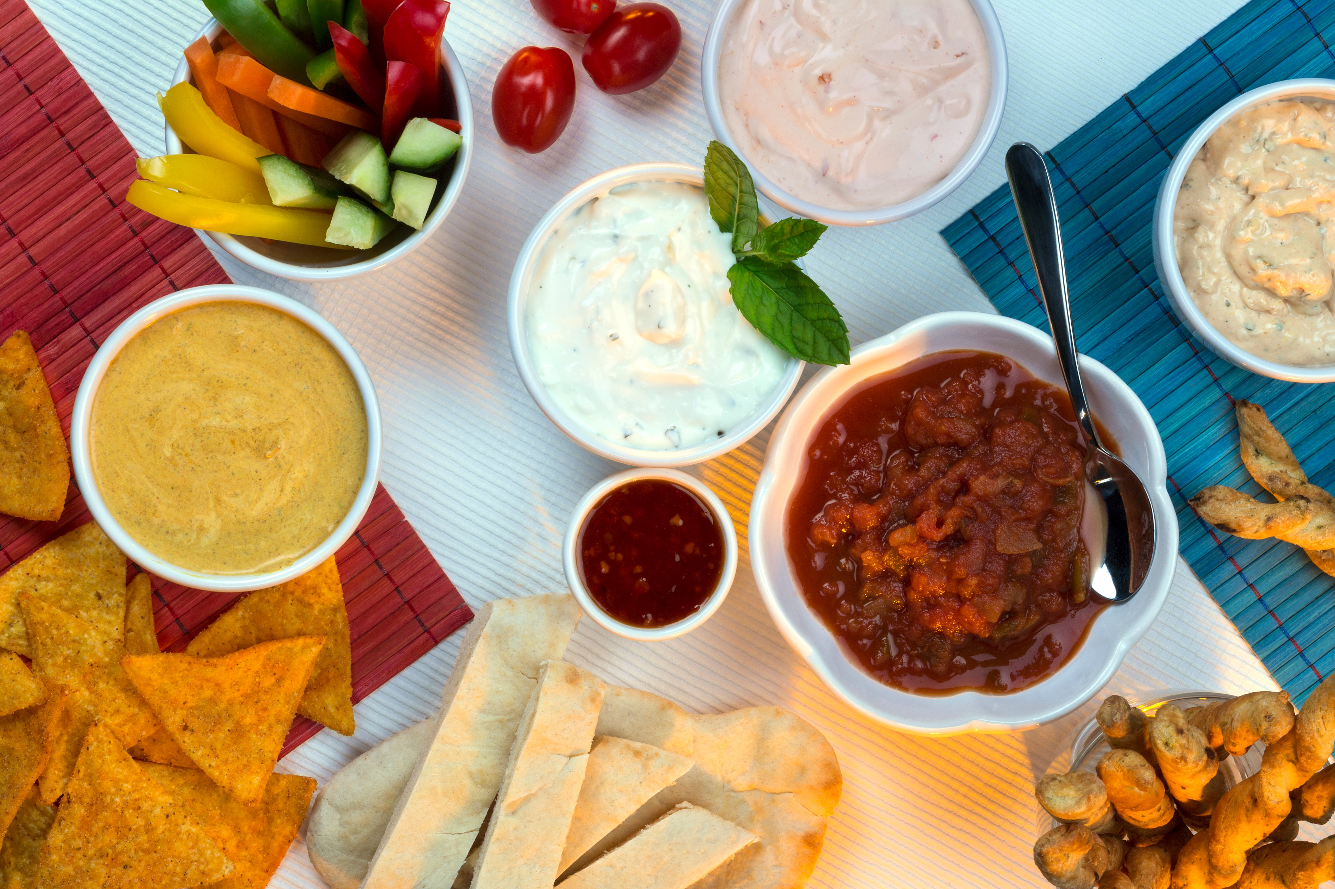 A collection of dips, breads, chips, and vegetables against a white table with red and blue placemats. 